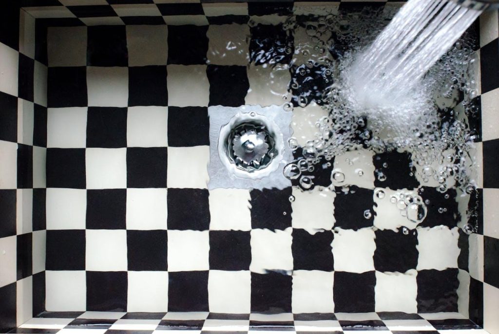 A shower head pouring water into a black and white checkered bathtub Description automatically generated